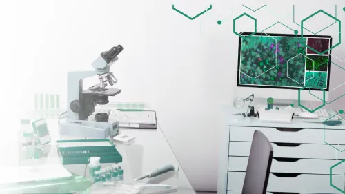 A laboratory desk with a microscope and a computer monitor displaying cellular imagery, overlaid with green geometric shapes.